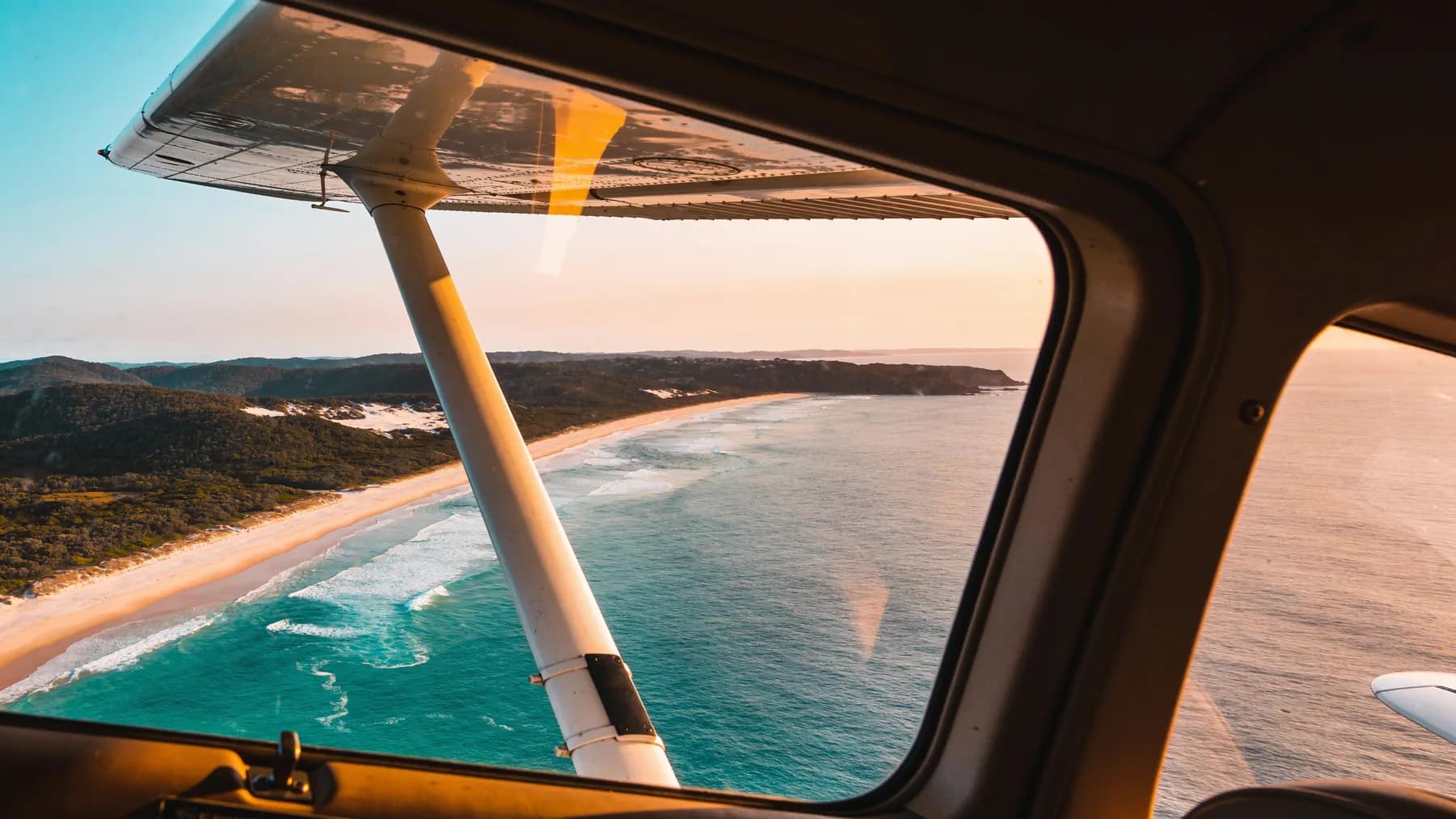 The view past the wing strut of a high-wing aeroplane over a turquoise Australian beach.
